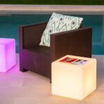 Lampe cube LED rechargeable blanc chaud à poser près d'un fauteuil en résine tressée au bord de piscine