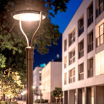 Lampadaire extérieur sur poteau métal gris, tête ronde plate avec bras incurvé et éclairage blanc