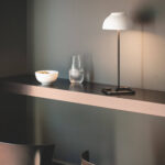 Lampe à poser blanche, pied fin en métal noir et abat-jour rond, posée sur tablette murale