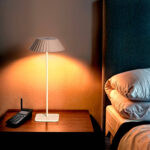 Lampe à poser blanche à abat-jour plissé, pied fin et base rectangulaire émettant une lumière chaude