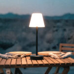 Lampe à poser anthracite, abat-jour blanc conique, pied fin et base circulaire, posée sur table extérieure en bois