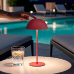 Lampe à poser portable rouge, abat-jour dôme sur tige fine, posée sur table extérieure près d'une piscine