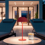 Lampe à poser rouge, pied fin et abat-jour rond sur table extérieure près de transats