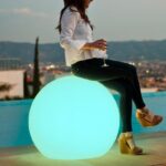 Sphère lumineuse extérieure turquoise, forme ronde, posée au sol et utilisée comme assise près d'une piscine