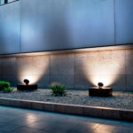 Spot extérieur noir sur socle rectangulaire, tête sphérique orientable projetant lumière chaude vers le mur