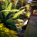 Projecteur extérieur au sol noir, boîtier métallique angulaire, lumière dirigée vers végétation pour éclairage d'accent